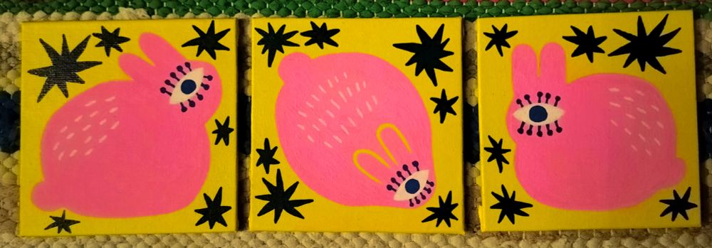 Photo of three small square paintings that are next to each other. All of them have bright yellow backgrounds and bright pink round bunnies with one single eye that has thick eyelashes. There are very dark blue stars in the background.