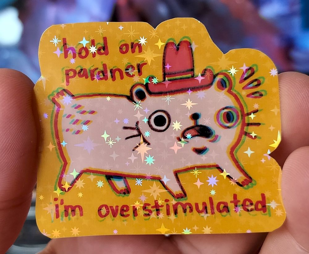 Photo of a yellow glittery sticker with some kind of rodent that resembles a hamster. It looks like it is in distress, standing up and sweating. There is a red cowboy hat on the head. Text around the animal reads "Hold on pardner, I'm overstimulated" There is slight chromatic aberration effect on the sides, really hitting the sentence home