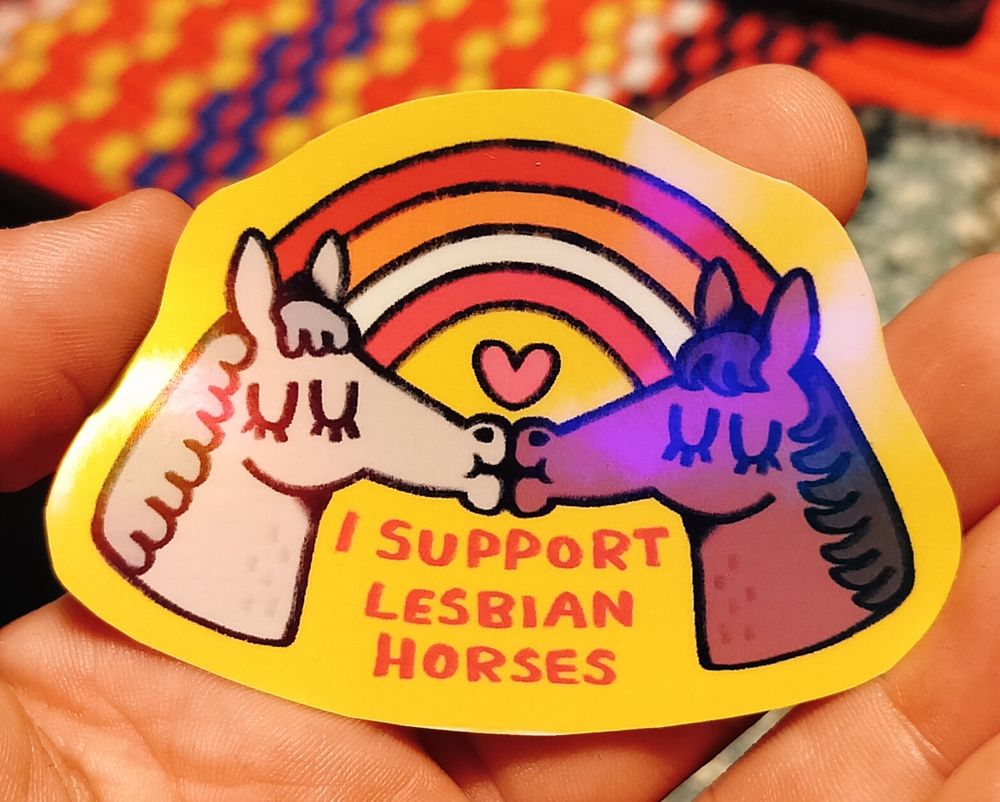 Photo of a sticker with two horses, one grey and one brown. They are kissing on a bright yellow background where there is also lesbian flag colored rainbow. There is a pink heart below their noses and darker pink text "I SUPPORT LESBIAN HORSES" under the heads.