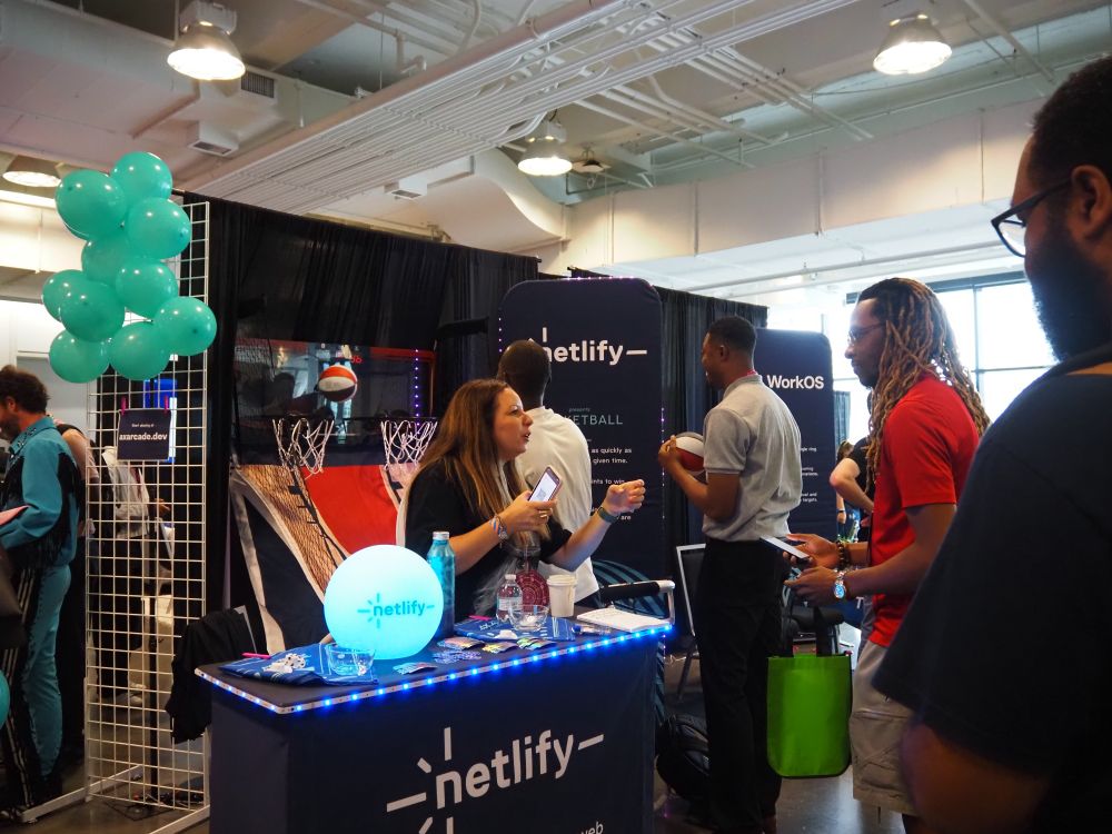An interactive event space featuring a Netlify booth with attendees talking and engaging. The booth includes teal balloons, a glowing Netlify orb, branded materials, and an arcade-style basketball game.