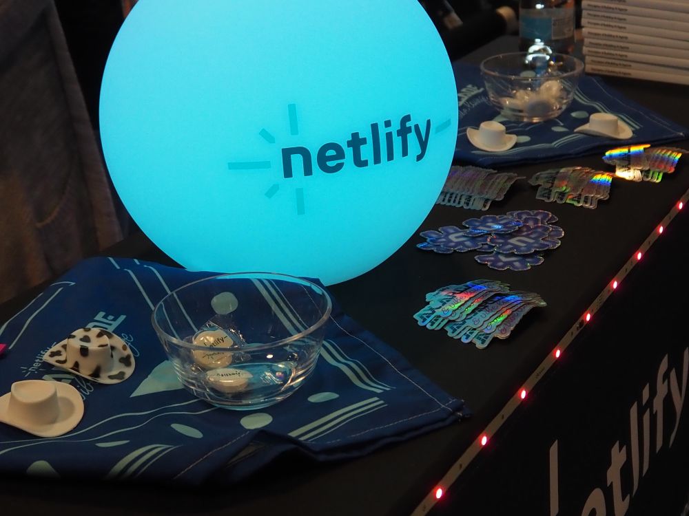 A close-up of a Netlify event booth table featuring a glowing blue orb with the Netlify logo, assorted shiny holographic stickers, small branded cowboy hats, clear bowls with pins, and blue bandanas arranged neatly.