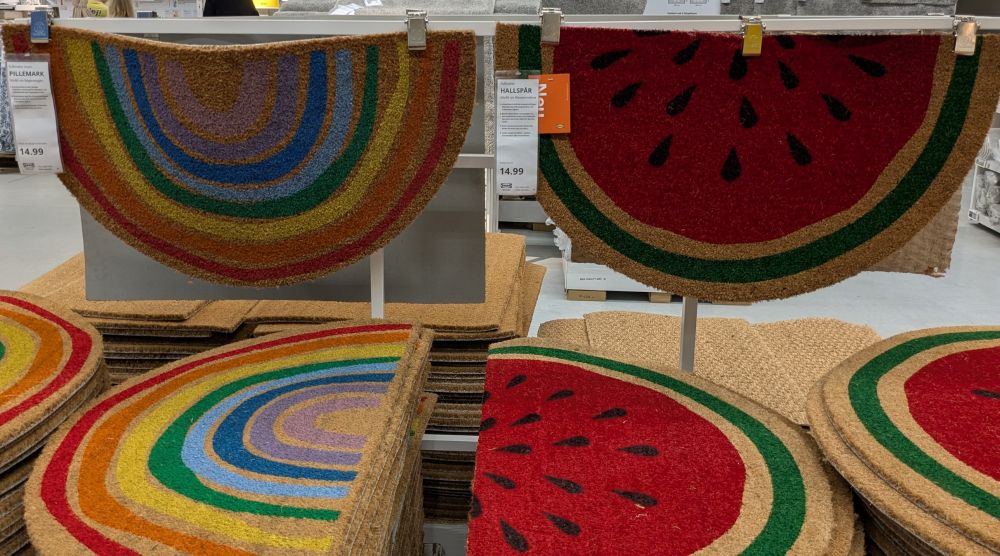 Mehrere Stapel Fußabtrittmatten bei Ikea. Rechts als Halbkreis eine aufgeschnittene Melone, links ein Regenbogen.