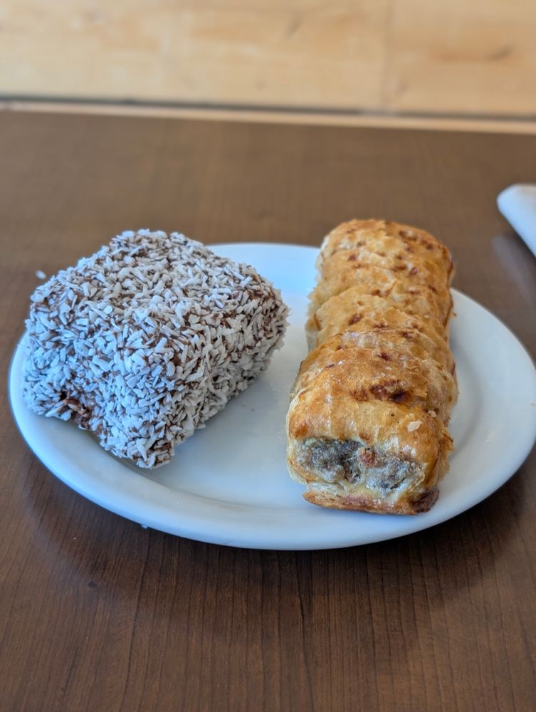 A white plate on a table with a lamington and a sausage roll on the plate.