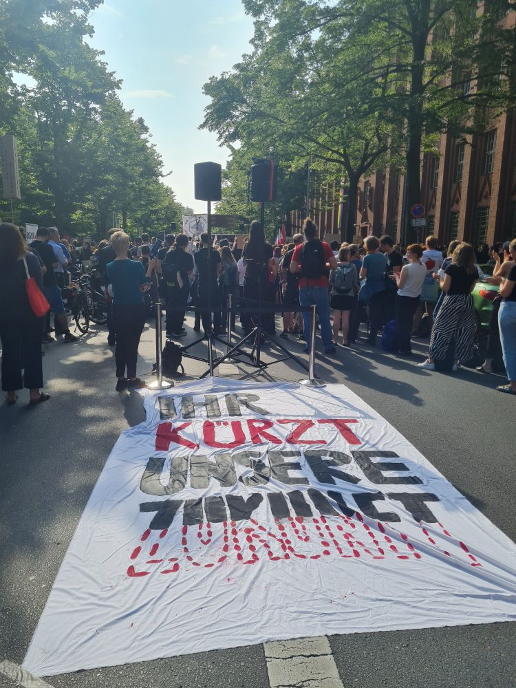 Auf dem Boden liegendes weißes Banner, darauf in Schwarz und Rot: "Ihr kürzt unsere Zukunft." Dahinter Kundgebungsteilnehmende. 