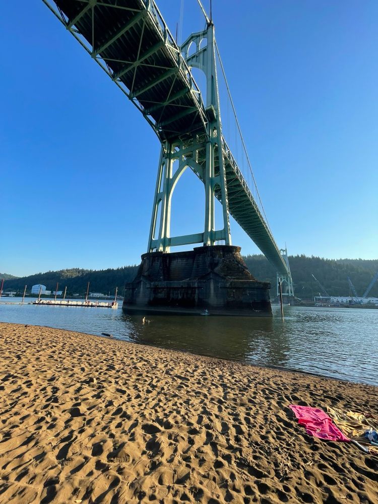 A photograph of the St. John’s Bridge and the beachfront area below. The beach is golden tan and Sandy, and the water is a murky green blue. There are two towels on the right side of the beach. In the water on the left side of the image you can see a white floating dock and people swimming at the dock. There are tree covered hill sides on the far side of the river With the setting sun reflecting off of them. The sky is blue with a yellow haze on the right side as the sun goes down.,