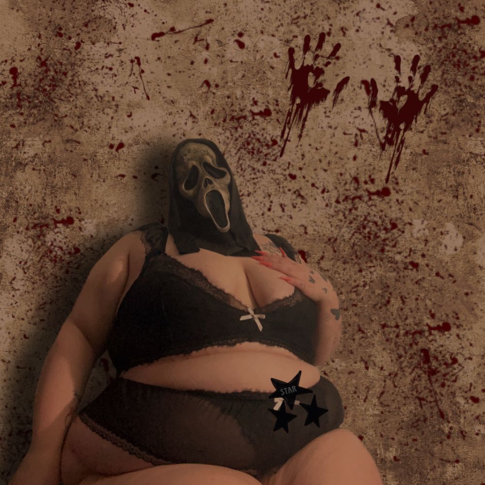 star in a black two piece sitting down a ghostface mask on and she has one hand resting on her tits 