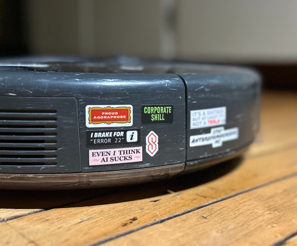 The rear bumper of a Roomba with little bumper stickers like 'Proud Agoraphobe' 'I brake for "Error 22"' 'Even I think AI sucks' and the cool S shape