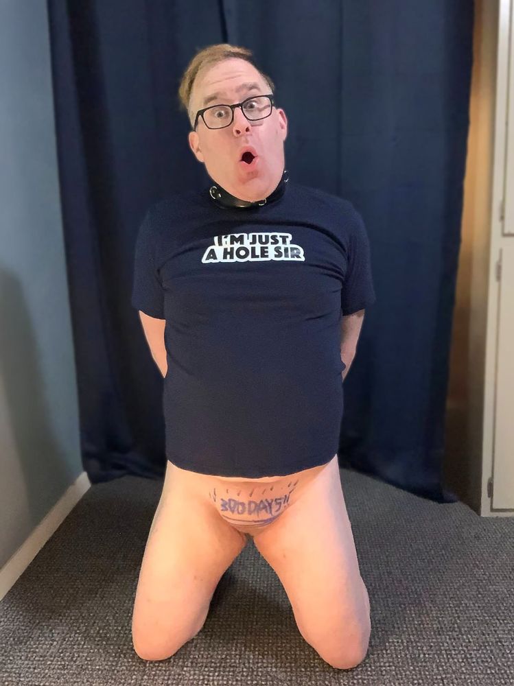 A nullo, a man without a penis and testicles, kneeling with his mouth open in a hole. His shirt says “I’m just a hole Sir” and he is wearing a leather collar. The text “300 Days” is written on his crotch. 