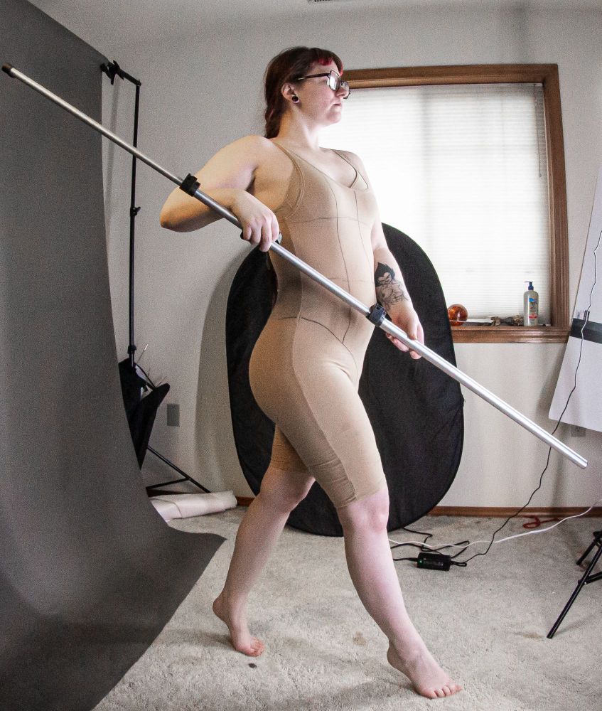 A photo from the side of a white person (Nyx Naia Stock) wearing a beige bodysuit and posing proudly with a long metal rod, holding it as though it were a staff.