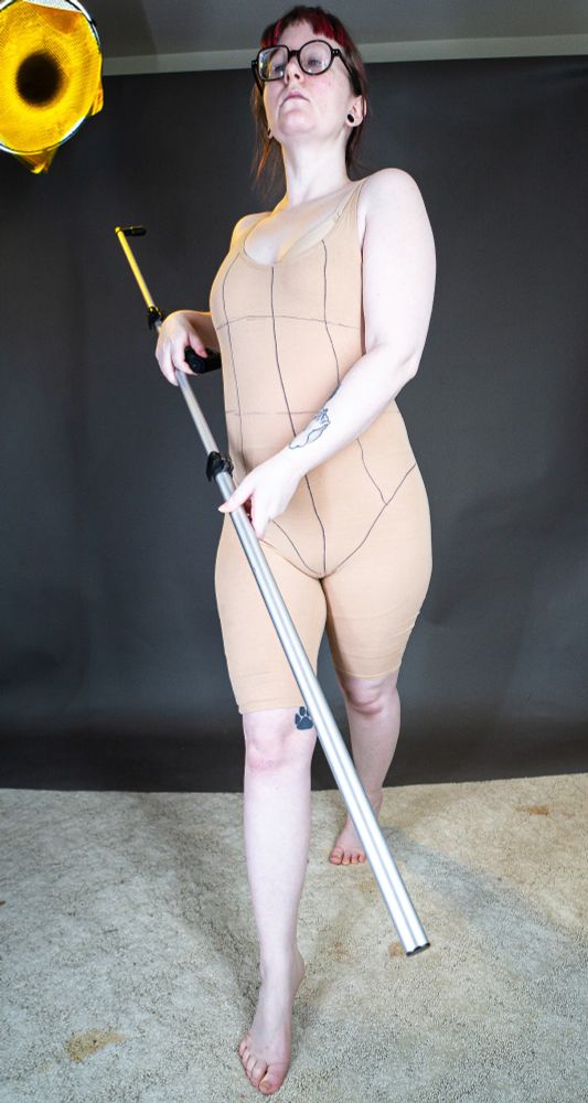 A photo from the front of a white person (Nyx Naia Stock) wearing a beige bodysuit and posing proudly with a long metal rod, holding it as though it were a staff.