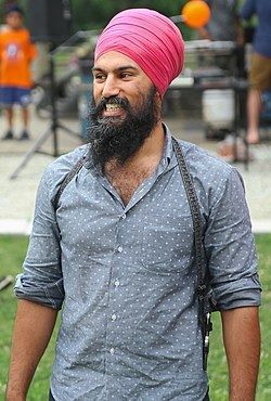 Jagmeet Sing, a Sikh man, is smiling in a park. He's wearing a bright pink turban with a gray shirt and has a long, curly beard.