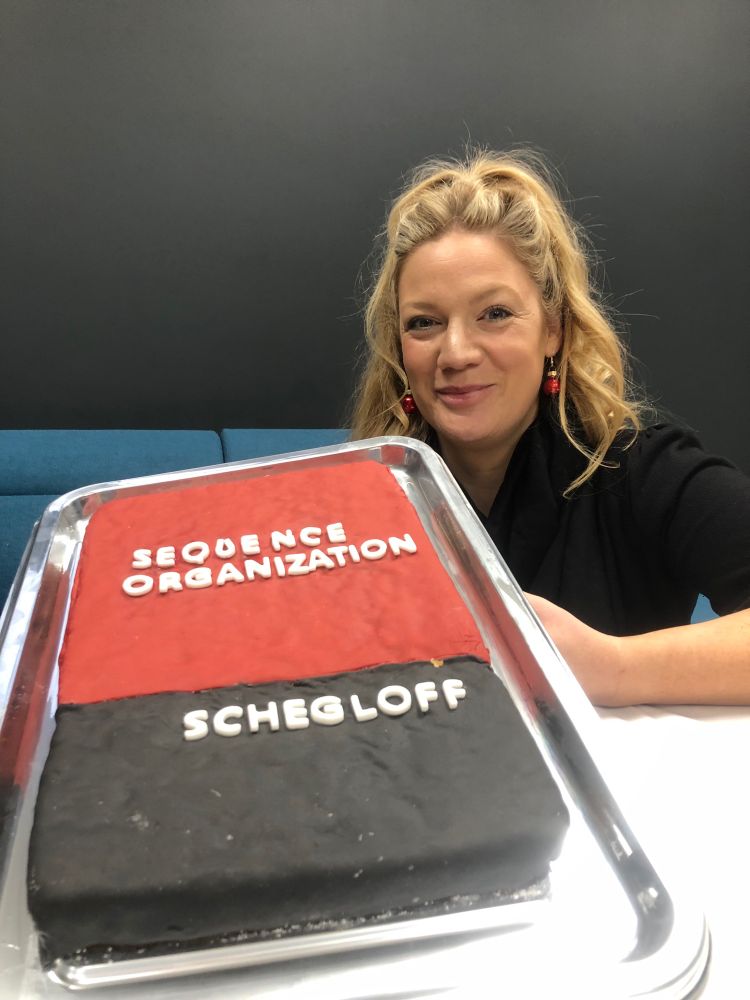 Red and white iced cake with the text “sequence organisation” and “Schegloff” with Emma posing in the background 