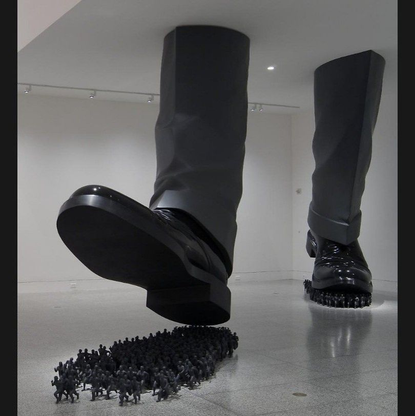 Sculpture 
D'immenses jambes avec pantalon de costume gris et souliers vernis s'apprêtent à écraser une foule minuscule sous leurs semelles

Credit : "Karma", oeuvre de l'artiste coréen Do Ho Suh