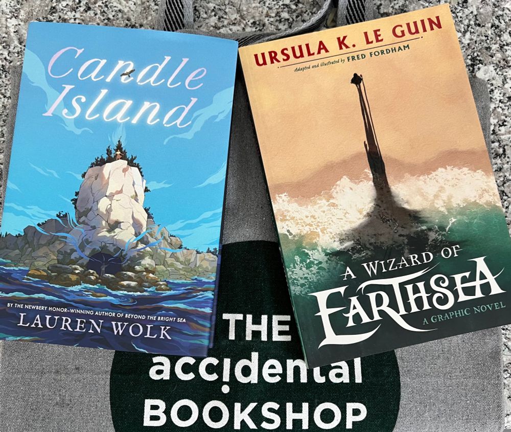 Grey tote bag with The Accidental Bookshop logo on it (white writing on green circular background).
Candle island by Lauren Wolk and A Wizard of Earthsea by Ursula Le Guin (graphic novel version) on top of the bag. 
