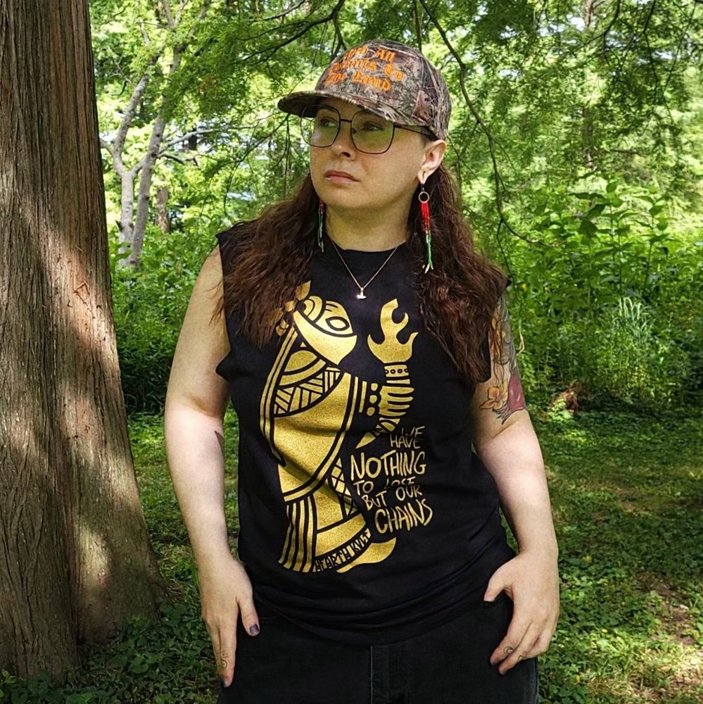 photo of me in a very green park. i'm wearing a black t-shirt with a golden-inked graphic, featuring a woman bearing a mead horn with fire spouting from it, her face covered, and the words "WE HAVE NOTHING TO LOSE BUT OUR CHAINS."