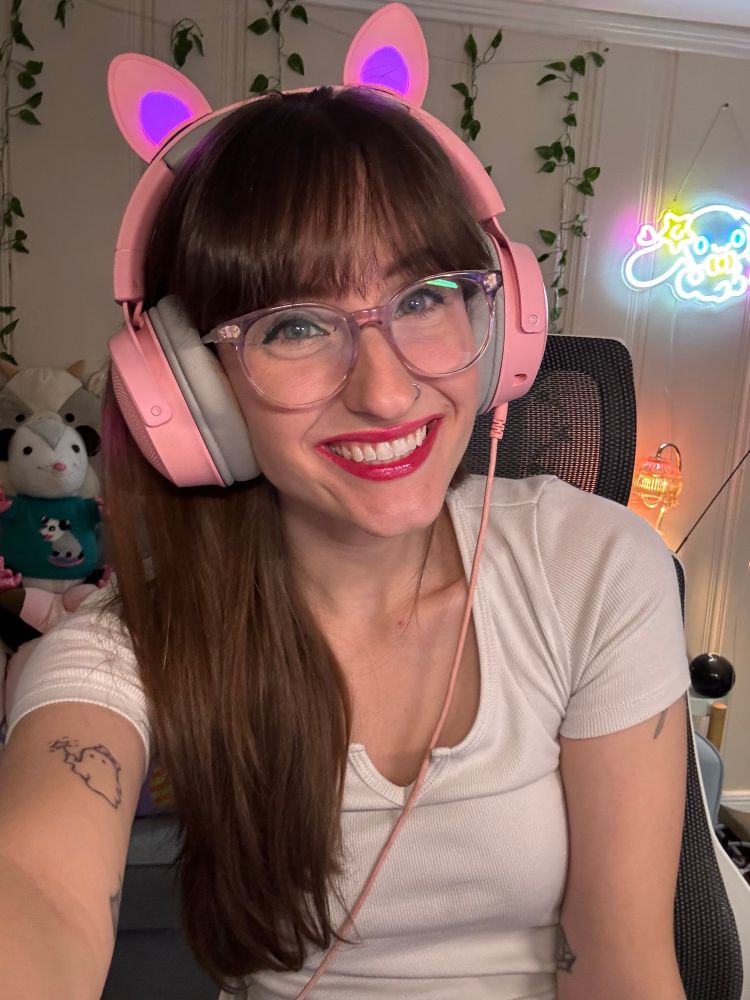 A selfie in which I'm sitting down and smiling at the camera. I'm a white woman with long brown hair and bangs and green eyes. I'm wearing a white t shirt, pink cat ear headphones, and clear frame glasses. 