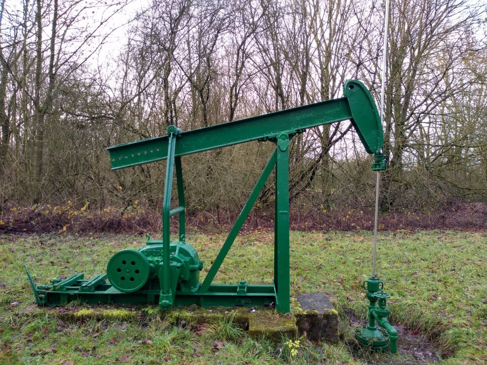 A photo of a small, old, green nodding donkey. It is not operational. It is overgrown with grass. There are trees in the background.