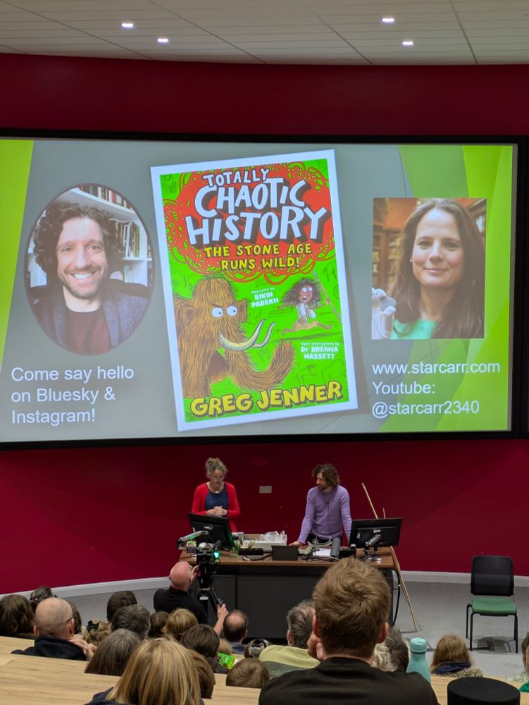 Photograph of a lecture. On the screen is a slide with a book titled 'Totally Chaotic history-the stone age runs wild' by Greg Jenner and Brenna Hassett. The book cover has a mammoth chasing a stone age person. On either side of the book image is a photo of Greg Jenner and Nicky Milner.