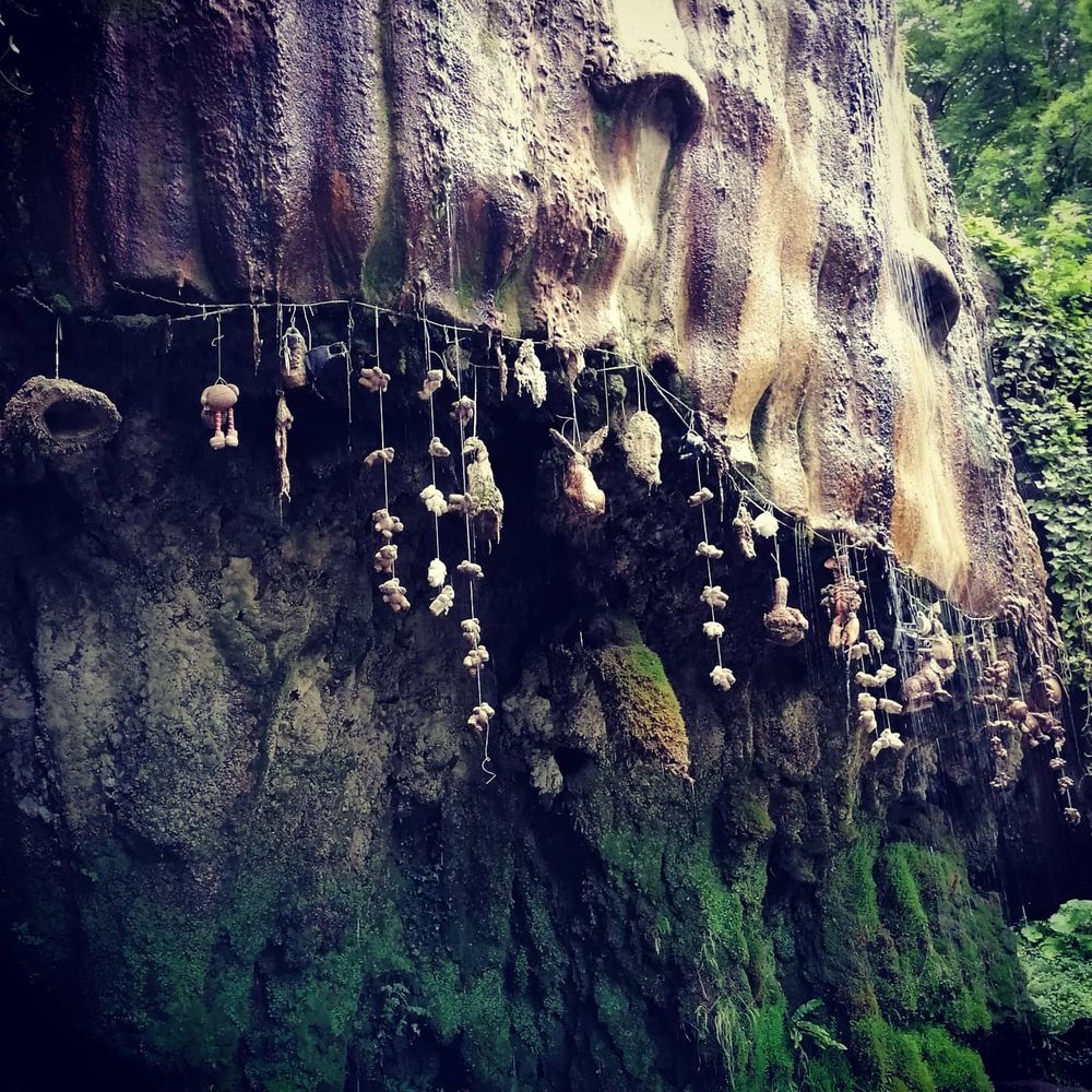 Photograph of a tufa waterfall. A small cliff is covered in tufa, and water drips down it. Under the overhang of the cliff are strings of objects - soft toys, hats etc - and they are getting covered in the calcium carbonate. 