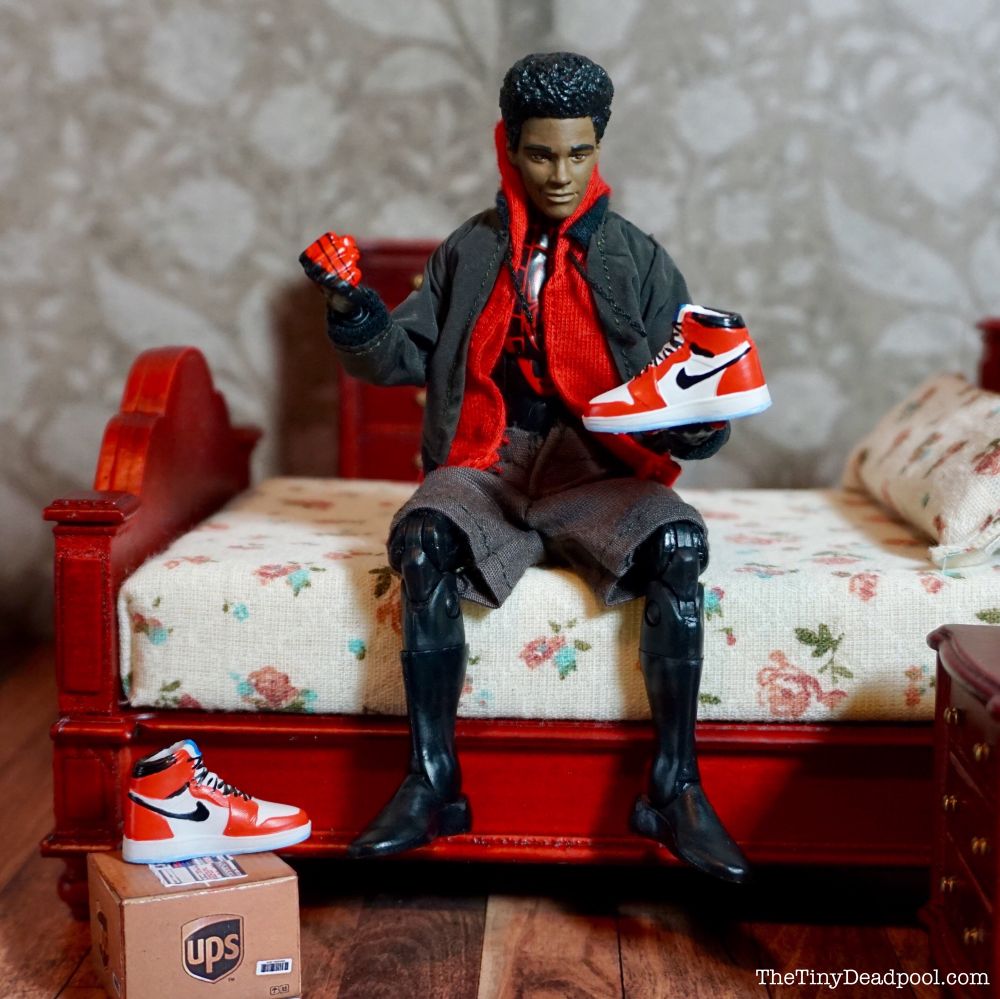 Teenage Miles Morales from Into the Spiderverse sits on a bed in his black colored spider man suit wearing a red hoodie and green coat over pumping his right fist while holding a white and red Nike Air jordan in his left hand with the ups box and matching shoe on the floor next to him - shot using a Hasbro marvel legends action figure.