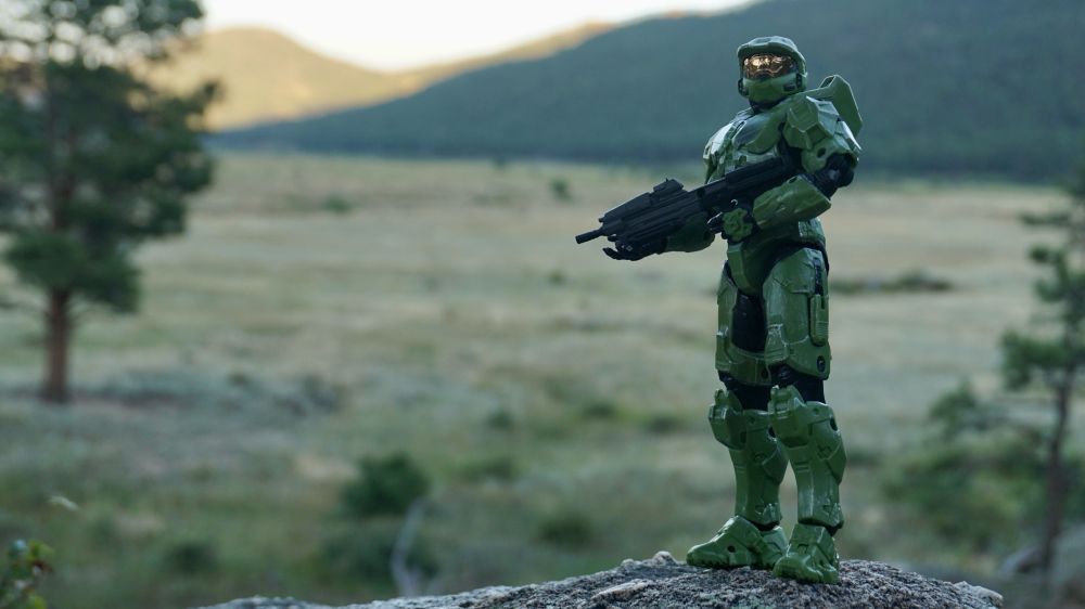 Halo’s master chief pauses on a rock outcropping near sunset with a field of grassland leading into foothills behind him