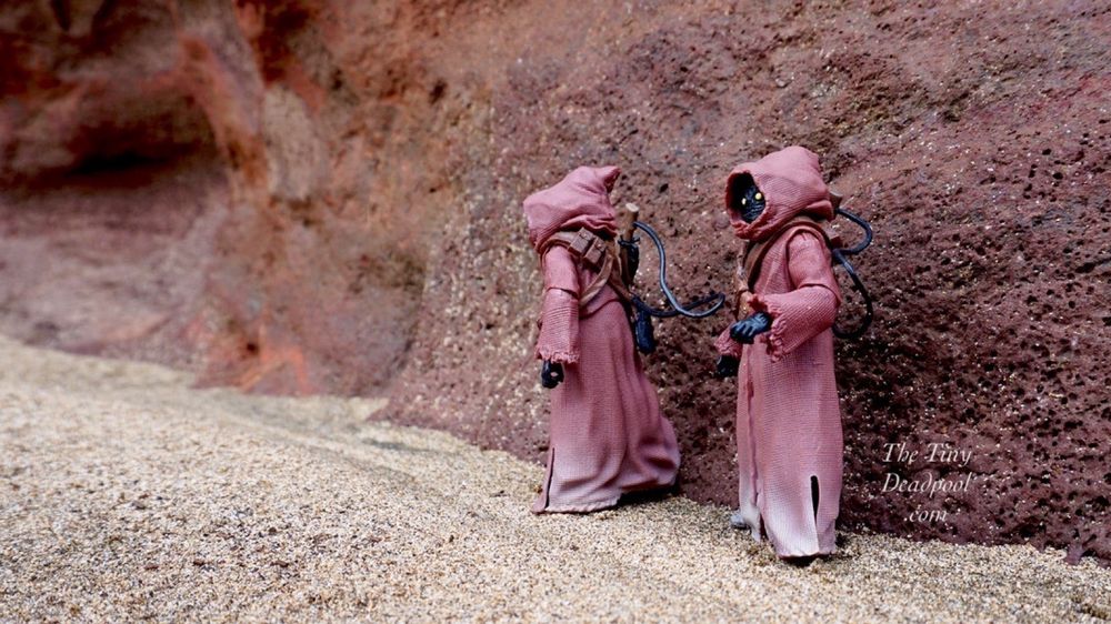 A Jawa walks uphill on the sand next to a red rock outcropping while his companion pauses to look to the distance wondering if he remembered to turn off water at home 