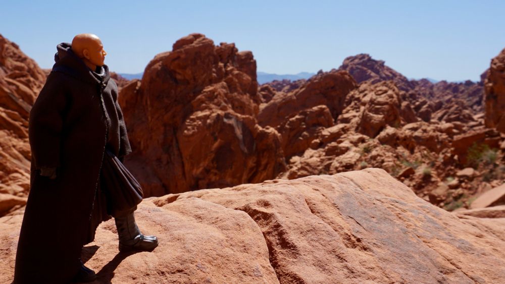 Boba Fett stands on an rust colored rocky shelf looking out into the hilly desert scape trying to decide where to go for lunch