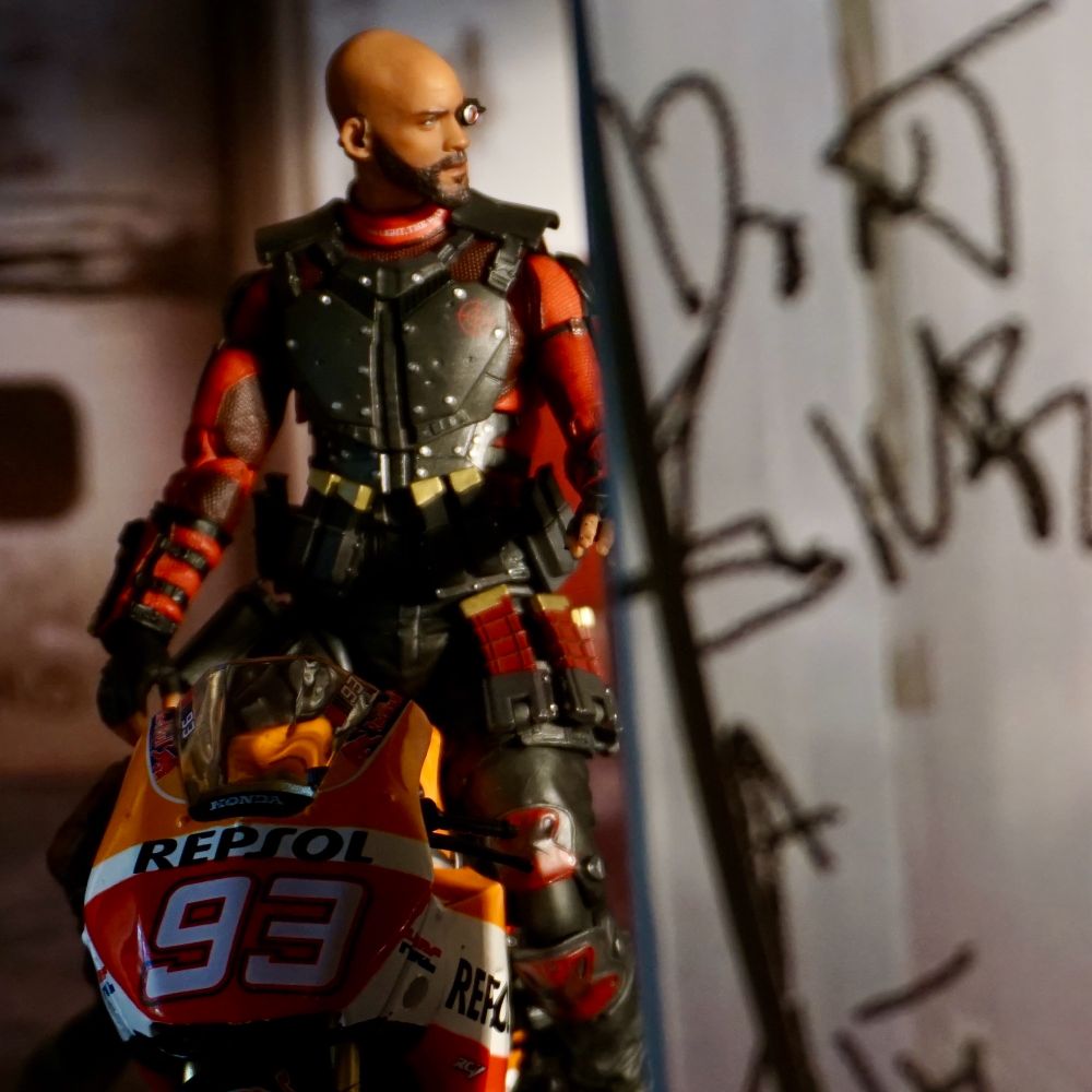 Deadshot sits astride a motorcycle looking off into the distance while stopped at a gas station 