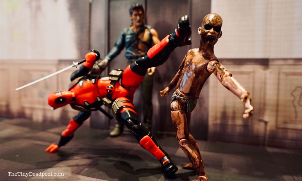Deadpool in an elevated horizontal position with his right hand and foot on the ground delivers a left footed kick to the head of a deadite as Ash from Evil Dead looks on in the background 