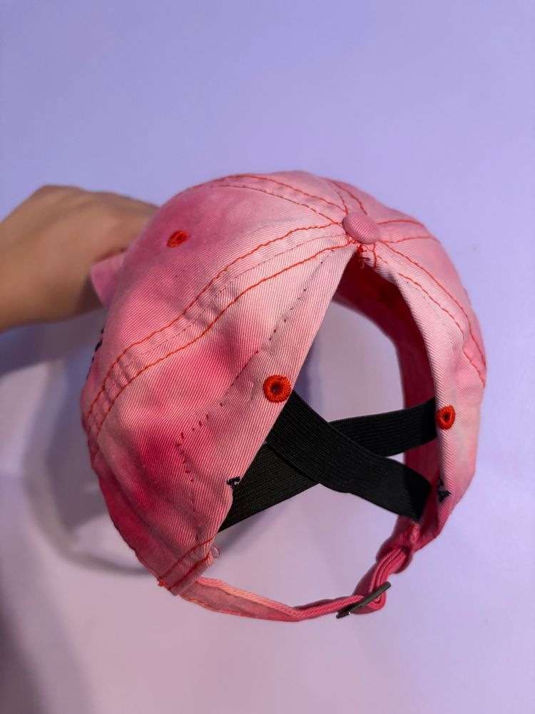 Pink baseball cap with the back opened for high ponytails. Two straps of elastic are crossed in the opening for structure.
