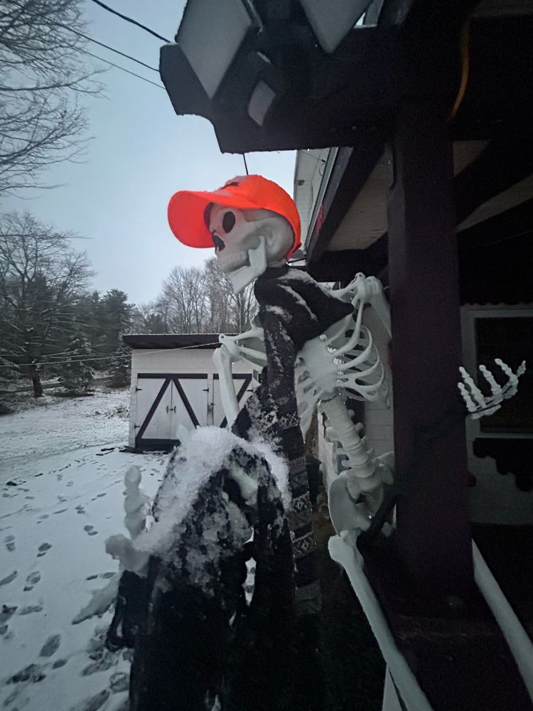 A skeleton with an orange hunter’s hat sits on a snowy porch. 🖤