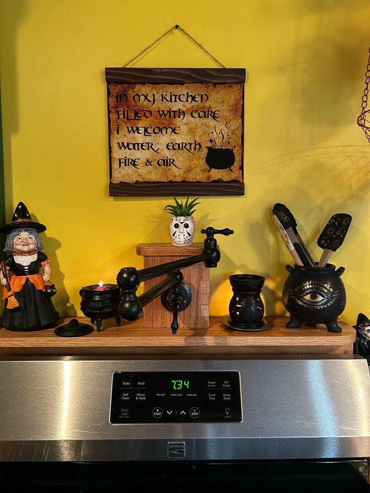 A sign above a stove reads “In my kitchen filled with care I welcome water, earth, fire & air” and sits above a mantle with cauldrons, candles and a witch. 