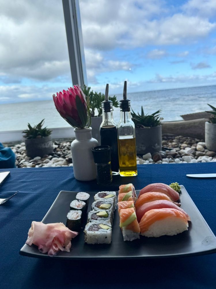 A photo of sushi platter with a Protea flower on the table. In the background some blue sky on a cloudy day over the sea. 
