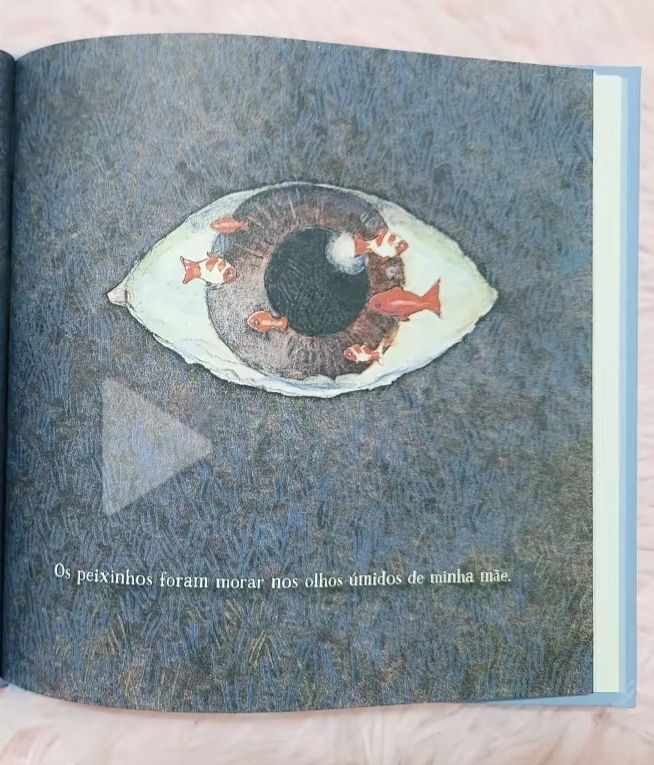 Página do livro Lá e Aqui de Odilon Moraes, o livro retrata um olho castanho com peixes dentro num fundo marítimo e a frase "Os Peixinhos foram morar nos olhos úmidos de minha mãe"