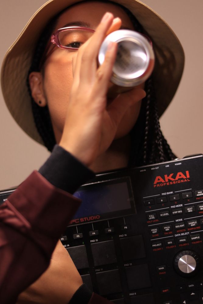Girl with skinny braids, wearing a beige bucket hat, drinking a beer and holding an electronic drum pad by AKAI 