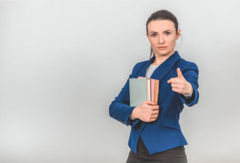 Feminine presenting person wearing a blue blazer and holding nondescript books, gazing intently at the viewer and pointing their left index at a point somewhere above the viewer’s line of sight