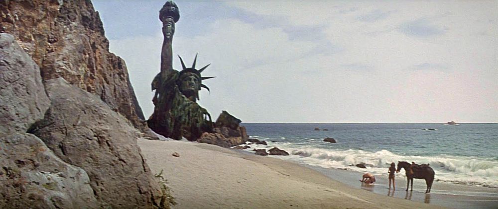 The top of the Statue of Liberty sticks out of the sand on a beach while a man drops to his knees in anguish next to a woman and a horse.