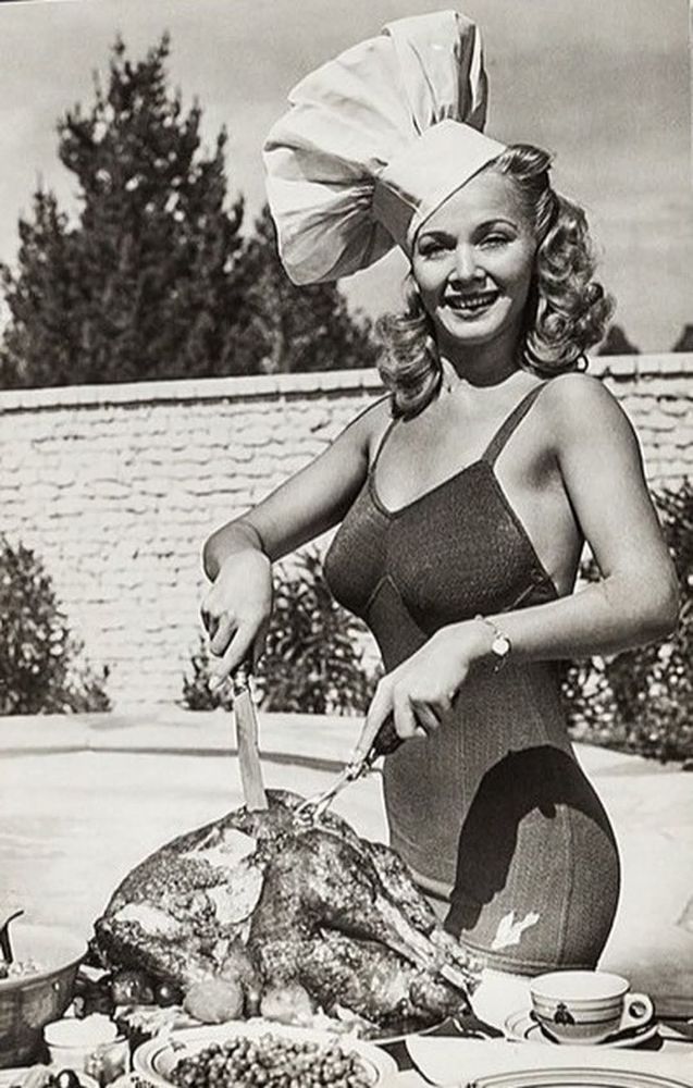A black and white photo of actress Carol Landis carving a very large turkey poolside while wearing what appears to be an exquisite one-piece bathing suit that hugs all of her ample curves. A chef's hat is perched at a jaunty angle upon her head as well.