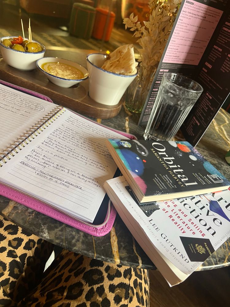 Cafe table surb writer’s notebook, a copy of Orbital, the booker prize winning novel and a copy of The Art of Creative Non Fiction. 