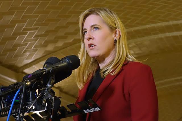 A deputada democrata Melissa Hortman, de Minnesota, fala ao microfone em uma coletiva de imprensa. Ela veste um blazer vermelho escuro e está diante de um fundo com teto dourado decorado. Diversos microfones estão posicionados à sua frente.