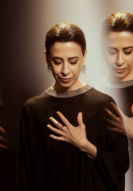 Fotografia de Fernanda Torres realizada para a Academia de Artes e Ciências Cinematográficas, organização responsável pelo Oscar. A atriz aparece em um retrato sofisticado, com expressão serena e olhar voltado para baixo. Ela usa um vestido preto elegante adornado com detalhes brilhantes na gola e brincos chamativos em formato de estrela. Sua mão repousa sobre o peito, transmitindo um gesto introspectivo. A imagem possui um efeito artístico de reflexos ou sobreposições sutis, conferindo profundidade à composição. A iluminação suave destaca sua figura em um fundo escuro e elegante.