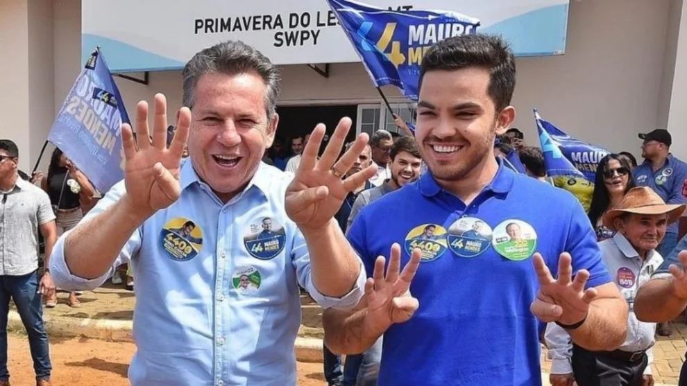 Mauro e o filho, sorridentes, posam lado a lado em frente a um prédio durante evento de campanha. Ambos usam botons e adesivos de candidatos, incluindo imagens de Jair Bolsonaro, e fazem com as mãos o gesto do número “44”.