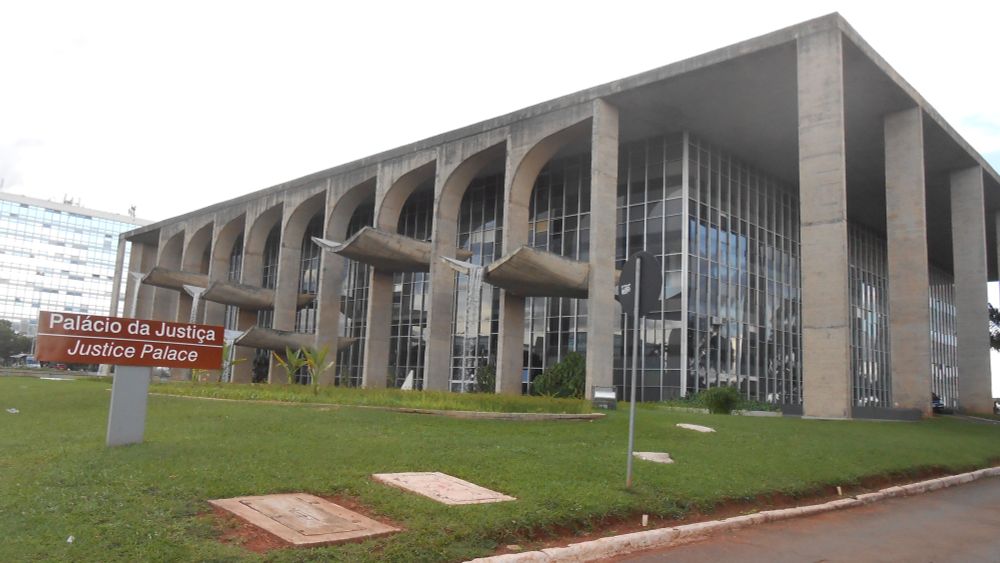 Foto do Palácio da Justiça, sede do Ministério da Justiça e Segurança Pública, localizado em Brasília (DF). O prédio é um ícone da arquitetura modernista de Oscar Niemeyer, com fachada composta por grandes colunas de concreto e vãos entre arcos marcantes. As estruturas horizontais que saem da fachada funcionam como espelhos d’água suspensos, com quedas d’água integradas ao paisagismo de Burle Marx. Na frente do edifício, há uma placa marrom com os dizeres “Palácio da Justiça” e a tradução em inglês “Justice Palace”. Ao fundo, parte de outro prédio espelhado do Eixo Monumental.