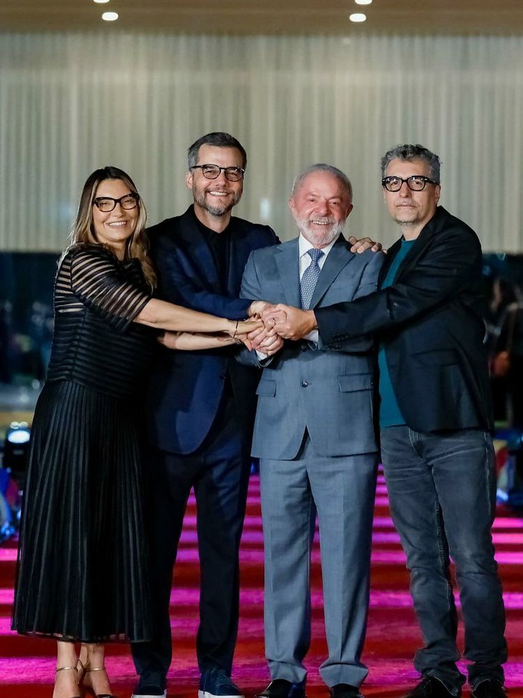 Foto mostra Janja, primeira-dama do Brasil, Wagner Moura, ator e diretor, Luiz Inácio Lula da Silva, presidente do Brasil, e Kleber Mendonça Filho, cineasta, posando juntos sobre um tapete vermelho. Eles estão lado a lado, sorrindo, e todos seguram as mãos uns dos outros no centro, em gesto de união. Janja usa vestido preto de mangas transparentes e saia plissada, Wagner Moura veste terno azul-escuro com camisa preta, Lula usa terno cinza com gravata azul de bolinhas e Kleber Mendonça está de blazer preto, camiseta verde-escura e calça jeans. Ao fundo, o ambiente é elegante, com cortinas claras e luzes no teto.
