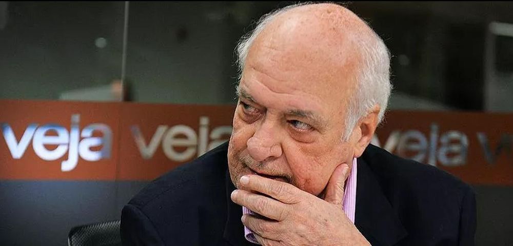 Foto mostra o jornalista J.R. Guzzo sentado em ambiente interno, com a mão parcialmente cobrindo a boca e o queixo, em um gesto típico de quem está refletindo ou avaliando algo com ceticismo. Ele veste blazer escuro e camisa social listrada em tom claro. Ao fundo, uma parede de vidro com o logo da revista Veja repetido em faixa vermelha, indicando que a imagem foi feita durante ou após uma entrevista ou participação em programa da redação. O olhar de Guzzo está levemente desviado para o lado, com expressão pensativa