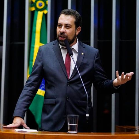 Foto mostra o deputado federal Guilherme Boulos (PSOL-SP) discursando no plenário da Câmara dos Deputados. Ele está de pé atrás da tribuna, com microfone frontal, usando terno escuro, camisa clara e gravata bordô. Boulos gesticula com a mão esquerda enquanto fala, com expressão firme e engajada. Ao fundo, a bandeira do Brasil está visível, reforçando o ambiente institucional.