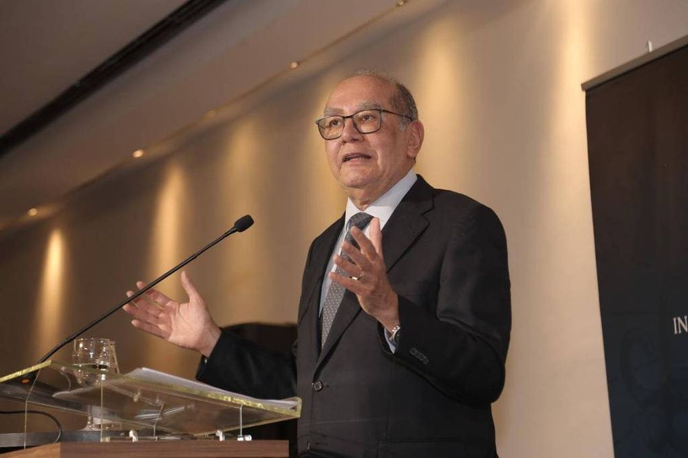 Foto mostra o ministro Gilmar Mendes, do Supremo Tribunal Federal (STF), durante uma palestra realizada em abril de 2025. Ele está de pé atrás de um púlpito transparente, vestindo terno escuro, camisa branca e gravata cinza com padrão discreto. Fala ao microfone com as mãos erguidas em gesto de explicação, com expressão atenta e tom de professor experiente. O fundo é neutro, com iluminação quente e um banner do evento parcialmente visível à direita. Imagem registrada por Jessica Boiczar e divulgada pela Avocar Comunicação e IASP.