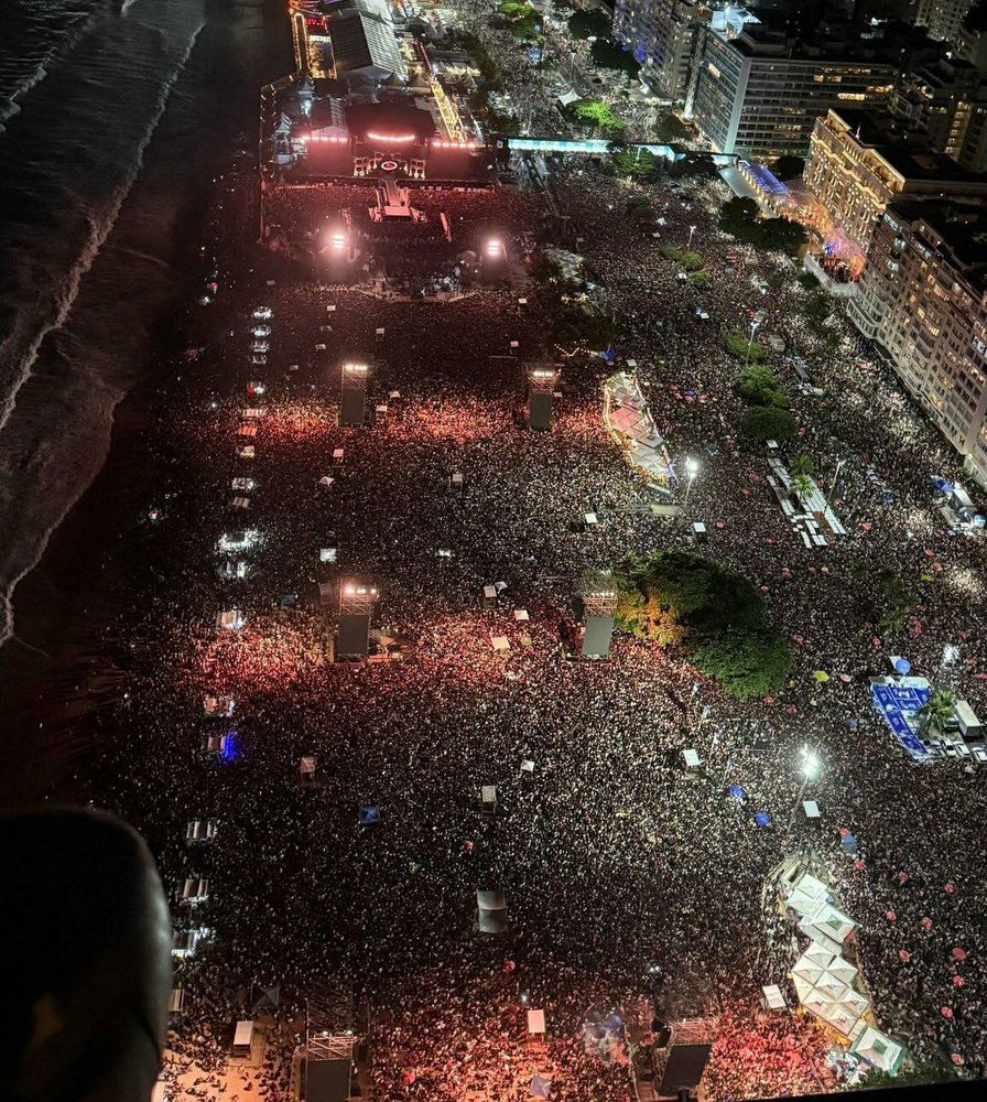 Foto aérea noturna mostra uma multidão compacta de mais de 2 milhões de pessoas reunidas na Praia de Copacabana, no Rio de Janeiro, diante de um grande palco iluminado. A imagem revela a orla à esquerda, o mar escuro, e os prédios iluminados à direita, com grande concentração de luzes e estruturas de som e segurança ao longo da praia.