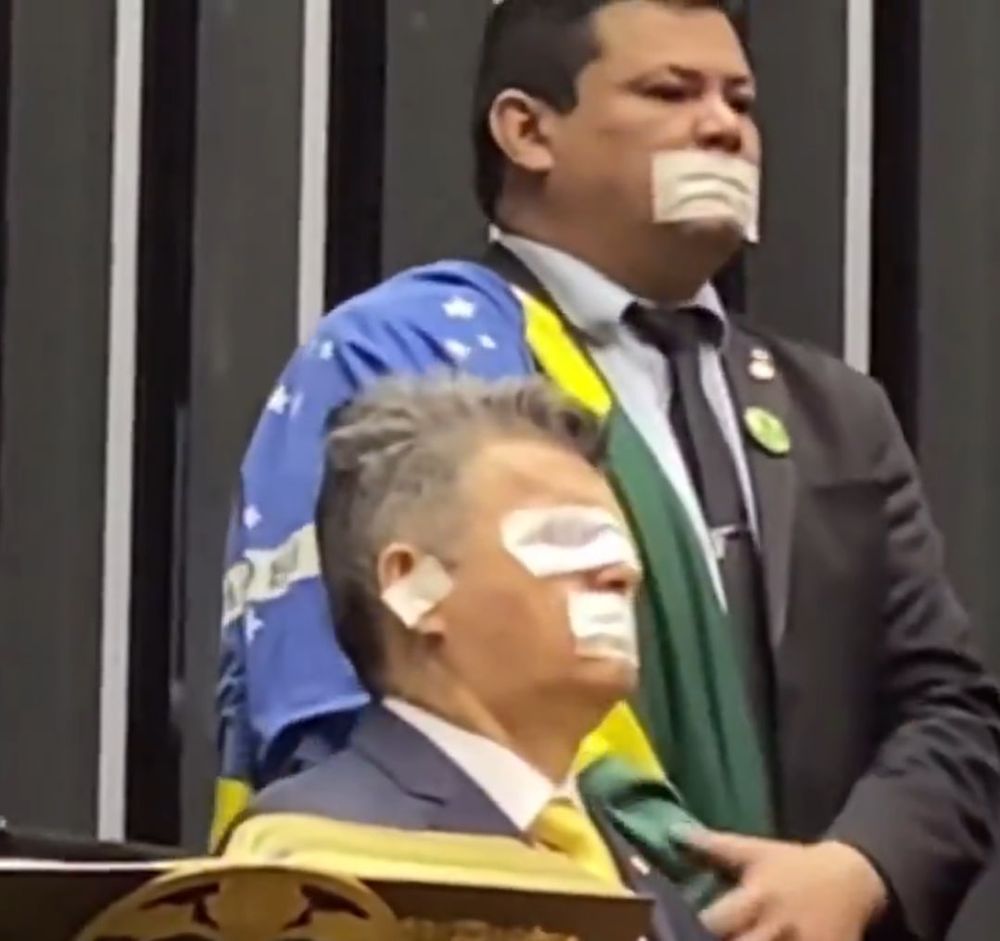 Dois deputados bolsonaristas aparecem na imagem dentro do plenário da Câmara dos Deputados, vestidos formalmente e com fitas adesivas coladas no rosto numa encenação grotesca de censura. O homem em pé está com a boca tapada e envolto em uma bandeira do Brasil, parecendo pronto para um cosplay de “mártir da liberdade”. Já o que está sentado exagera ainda mais: tem fita adesiva sobre a boca, olhos e ouvidos, como se encenasse o velho ditado dos três macacos — não vê, não ouve, não fala — só que com drama e patriotismo de loja de souvenir. Ambos usam terno escuro com gravata amarela, reforçando a identidade estética do bolsonarismo. A ação faz parte da tentativa de “sequestro simbólico” da Mesa Diretora da Câmara, feita por parlamentares da extrema direita em protesto contra decisões do STF e da presidência da Casa. Cena que mistura teatro político, delírio e TikTok.