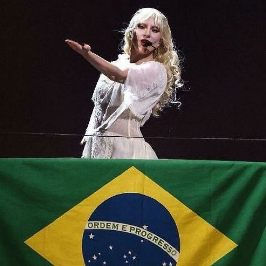 A cantora Lady Gaga se apresenta em um palco com fundo escuro, usando um vestido branco rendado e microfone de cabeça. Ela está atrás de uma bandeira do Brasil estendida à sua frente e gesticula com o braço direito estendido para o lado esquerdo.